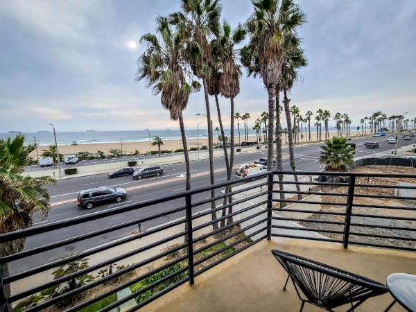 A beach is visible from a balcony with palm trees, a road, and cars. The scene extends to sand and ocean under a cloudy sky in the background.