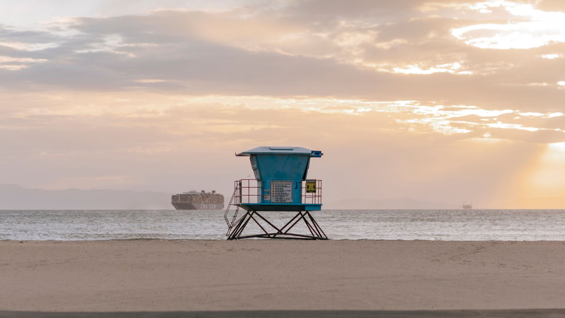 A beach with a lifeguard tower in the center, cargo ships in the distance, and a serene sunset sky.