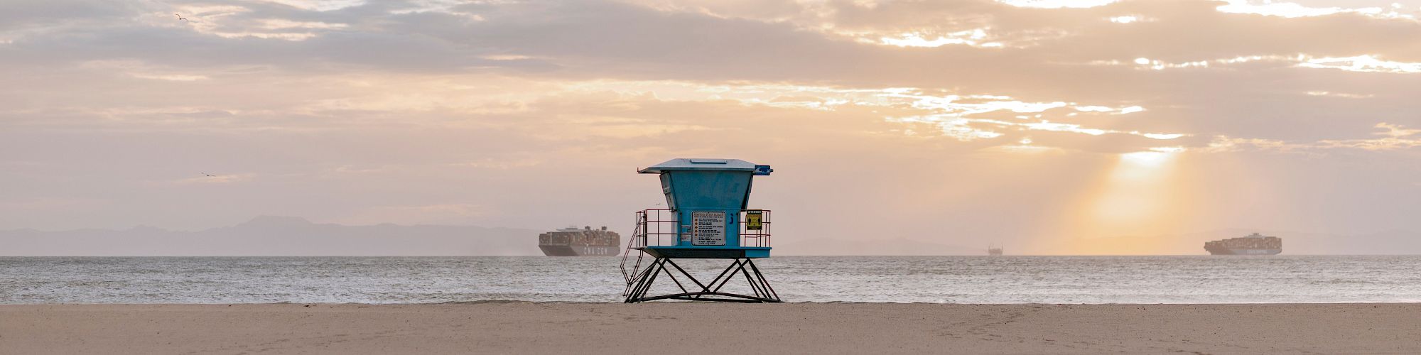 A beach with a lifeguard tower in the center, cargo ships in the distance, and a serene sunset sky.