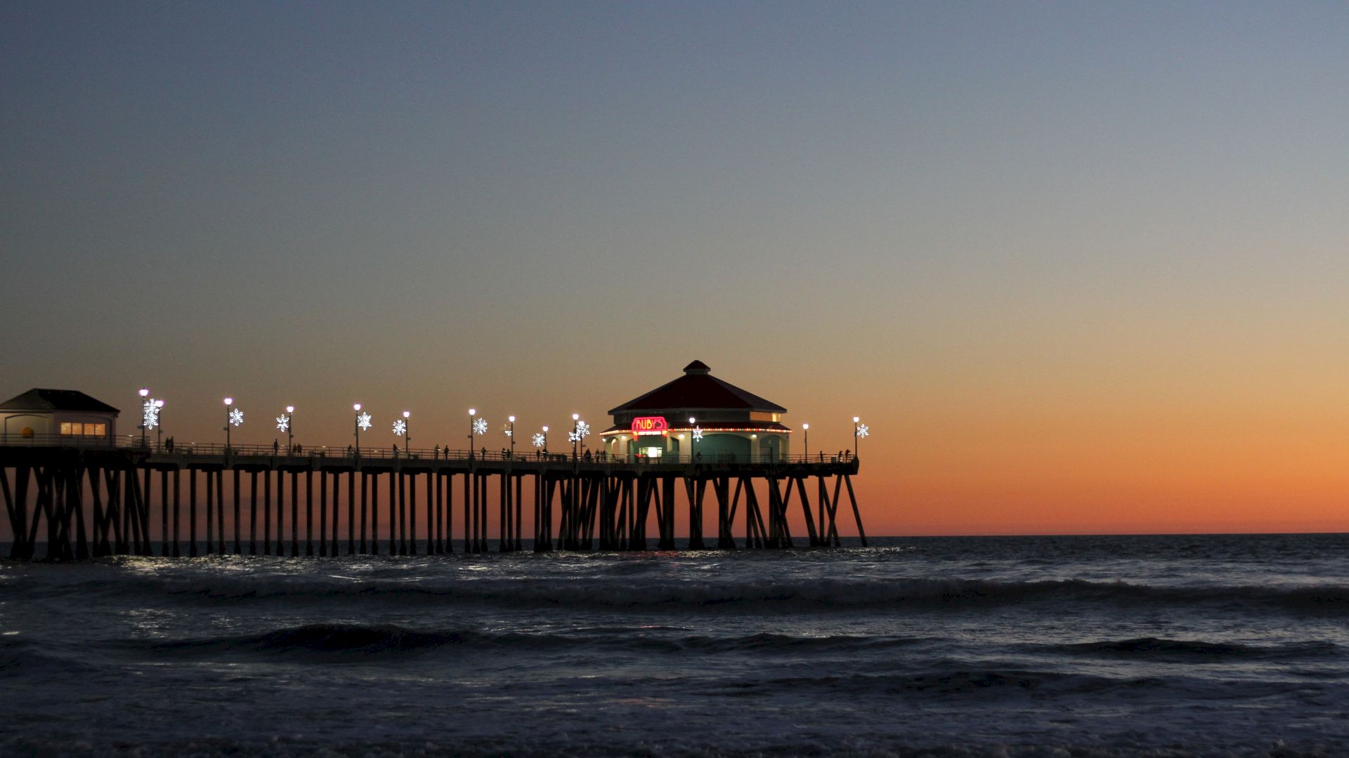 A pier extends into the ocean under a twilight sky, with lights illuminating the walkway and a building at the end of the pier.