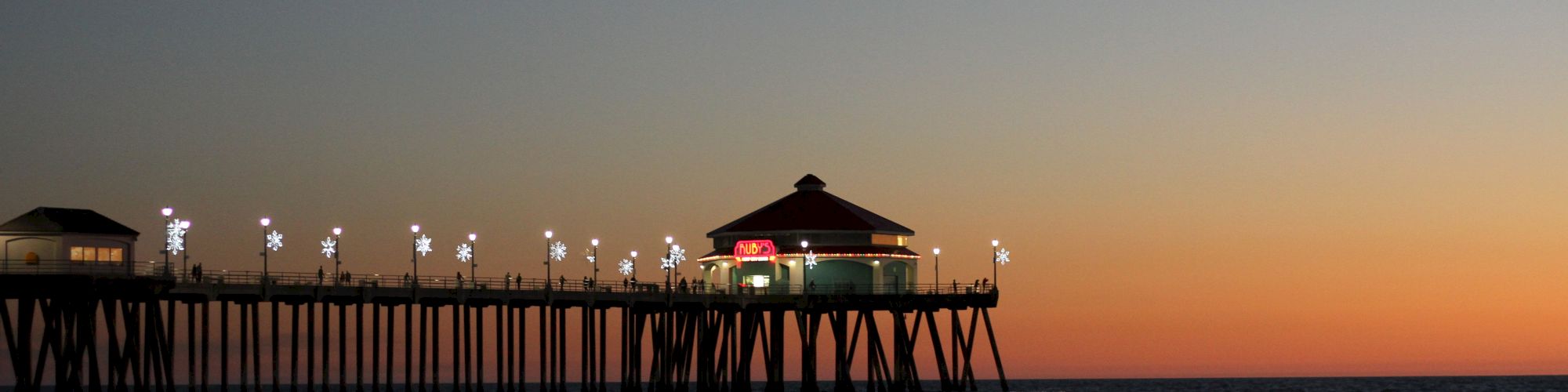 A pier extends into the ocean under a twilight sky, with lights illuminating the walkway and a building at the end of the pier.