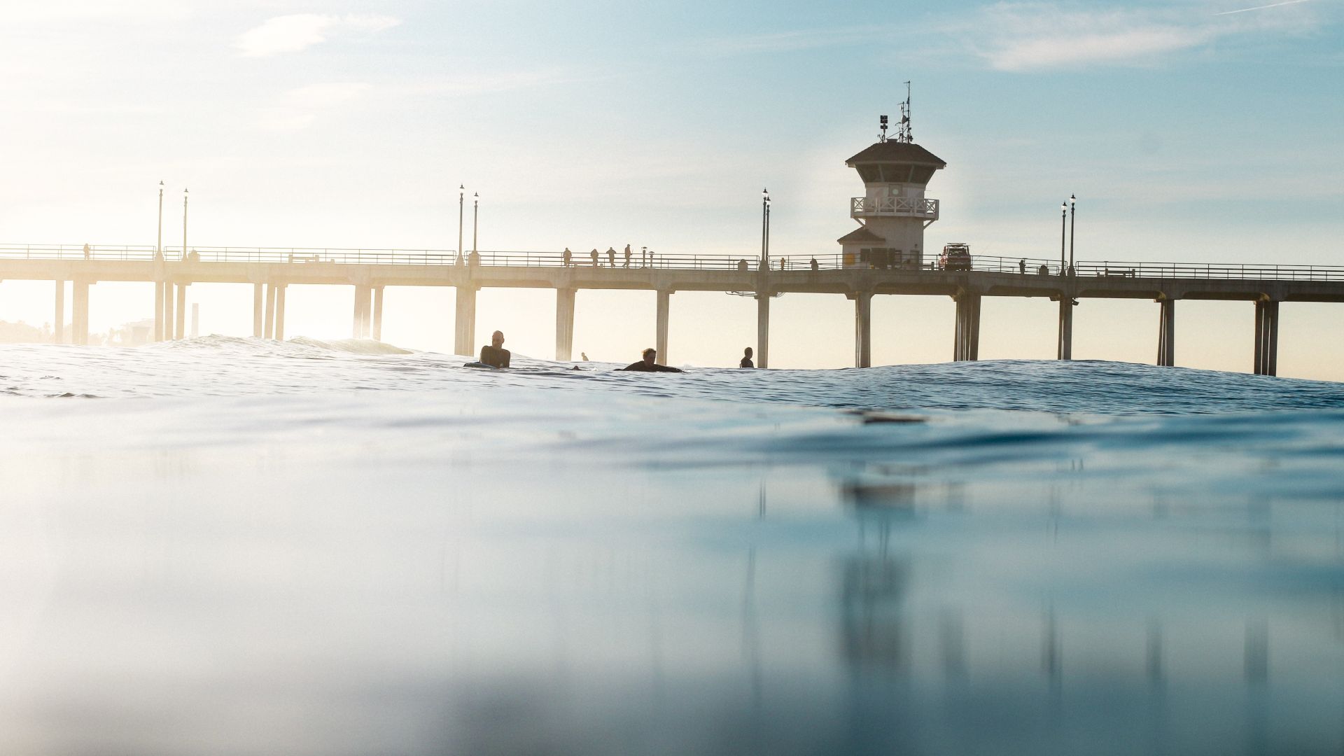 The image shows a pier extending over the ocean with people walking on it under a clear sky. The view is partially submerged in water, with surfers visible.