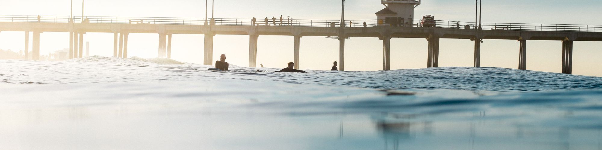 The image shows a pier extending over the ocean with people walking on it under a clear sky. The view is partially submerged in water, with surfers visible.