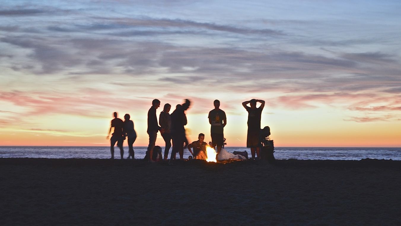 A group of people are gathered around a fire on a beach at sunset, silhouetted against a colorful sky, creating a serene and social scene.