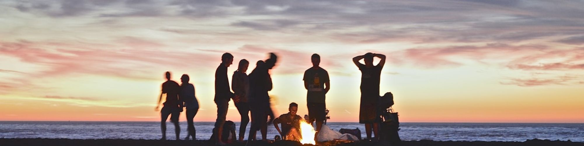 A group of people are gathered around a fire on a beach at sunset, silhouetted against a colorful sky, creating a serene and social scene.