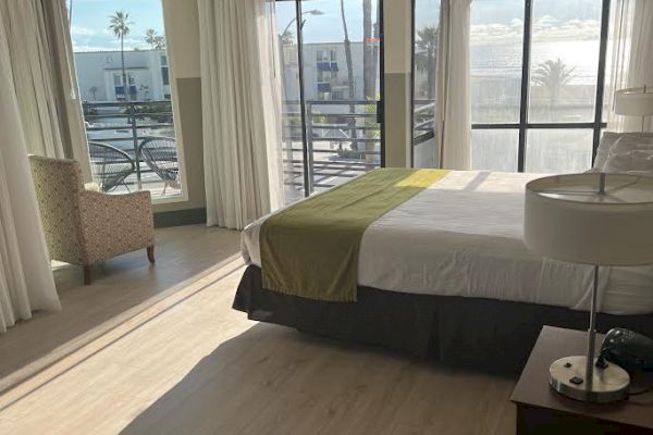 A brightly lit hotel room with large windows, a bed, bedside table with lamp, and a chair in the corner next to the window.