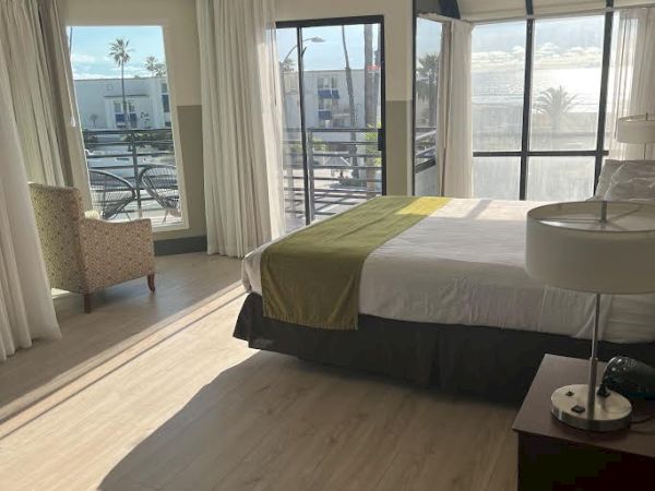 A brightly lit hotel room with large windows, a bed, bedside table with lamp, and a chair in the corner next to the window.