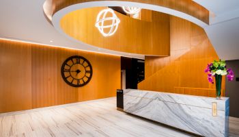 A modern lobby features a marble reception desk, wooden walls, a large wall clock, and a unique spiral staircase with spherical light fixtures.
