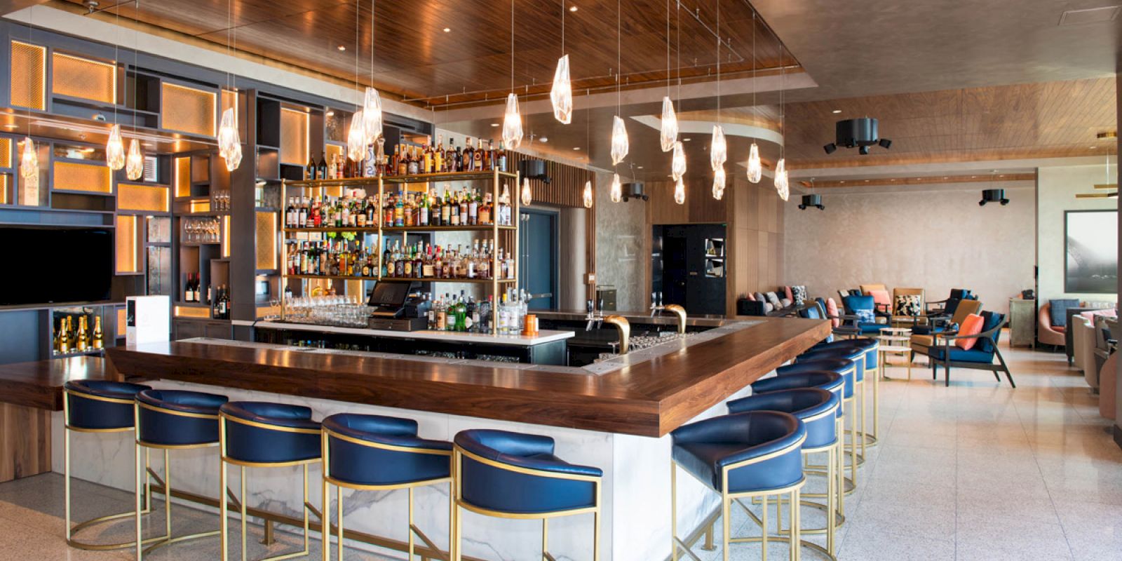 The image shows a modern bar with stylish seating, hanging pendant lights, and a well-stocked bar counter in a spacious, open setting.