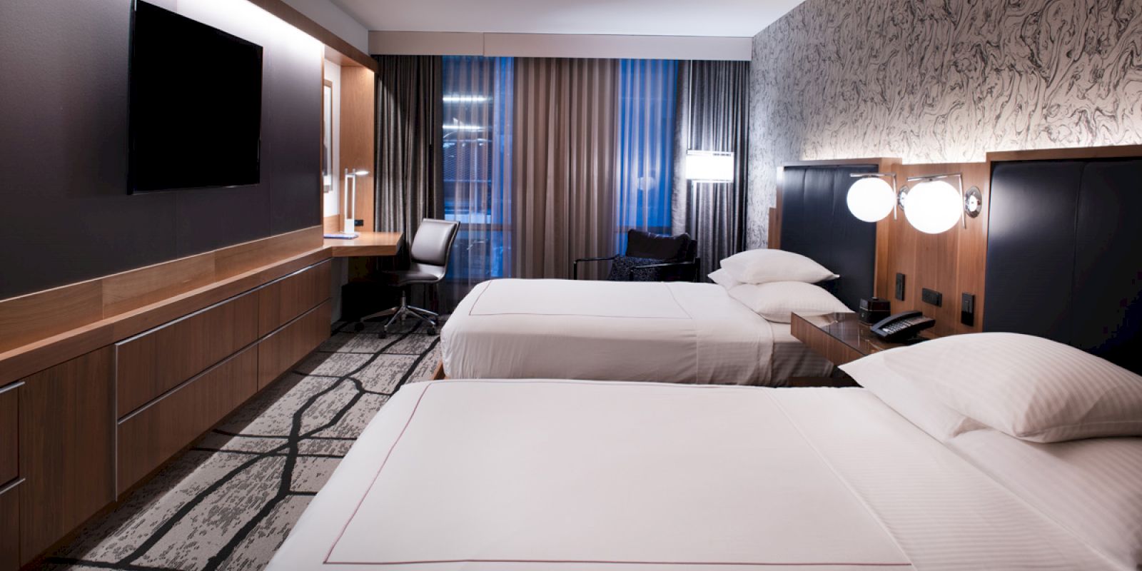 The image shows a modern hotel room with two double beds, a flat-screen TV, a desk with a chair, and contemporary decor and lighting.