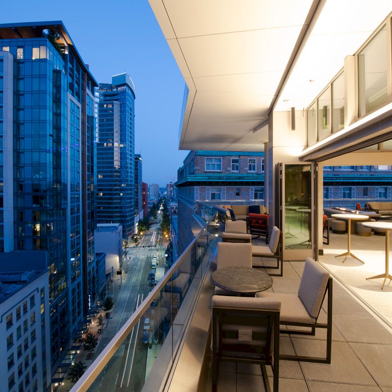 The image shows a modern outdoor terrace with seating, tables, and a cityscape view during dusk. The terrace overlooks high-rise buildings and a street.