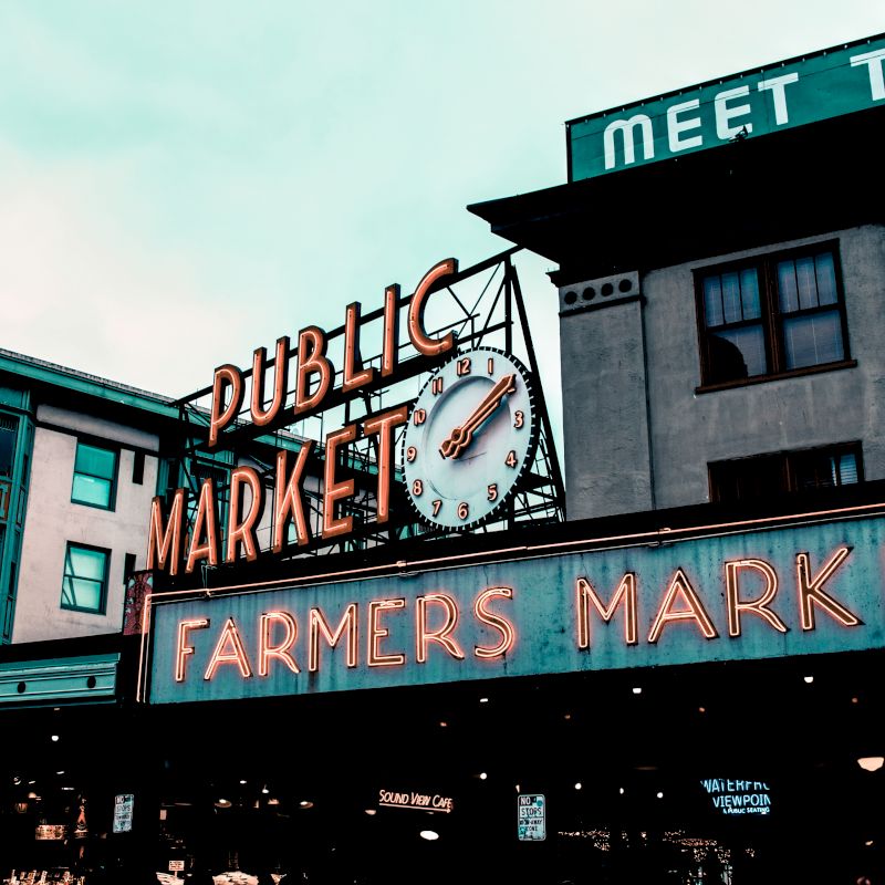 The image shows a famous public market with prominent "PUBLIC MARKET" and "FARMERS MARKET" neon signs on buildings, featuring a large clock.