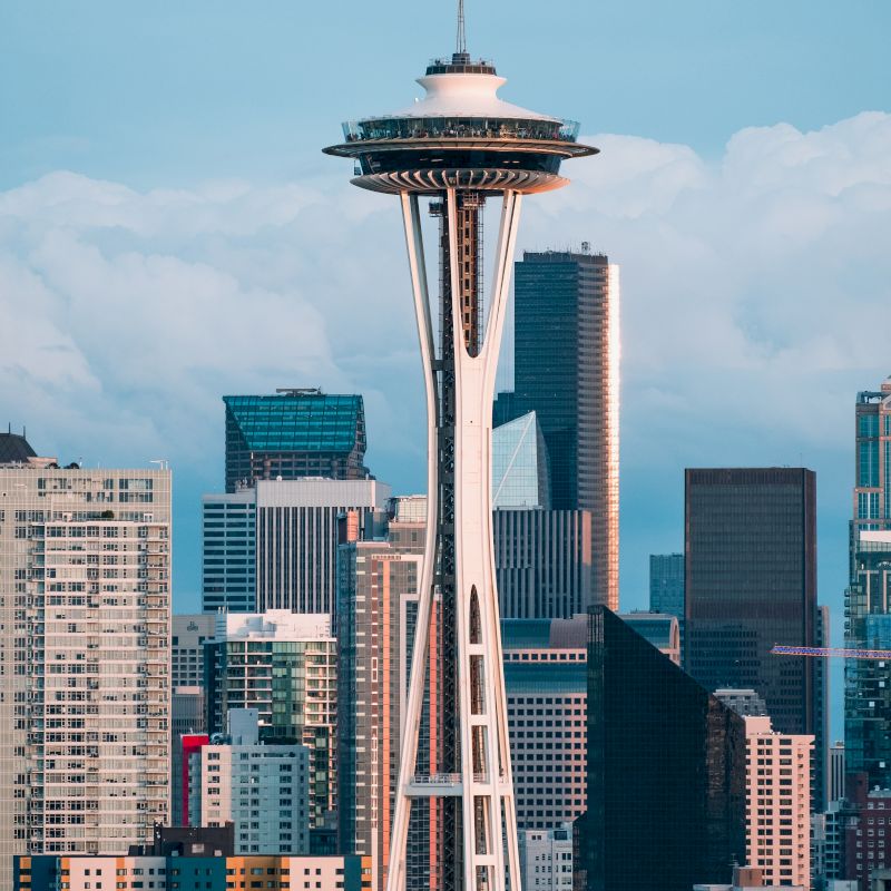 The image shows the Space Needle, an iconic observation tower in Seattle, Washington, with a backdrop of modern high-rise buildings and a cloudy sky.