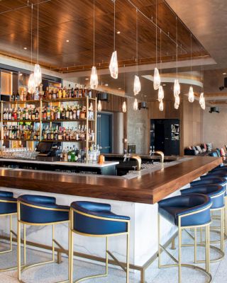 This image shows a modern bar with a wooden countertop, blue cushioned bar stools, hanging lights, and a well-stocked liquor shelf in a stylish lounge area.