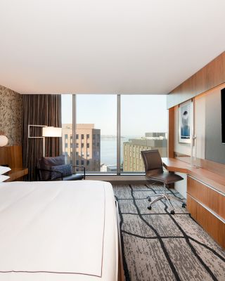 A modern hotel room with a large bed, TV, desk, chair, and a window offering a city view.