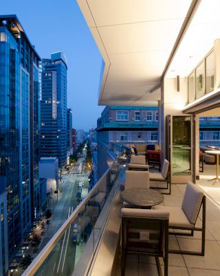 A modern, furnished balcony with tables and chairs overlooks a city street lined with tall buildings at dusk.