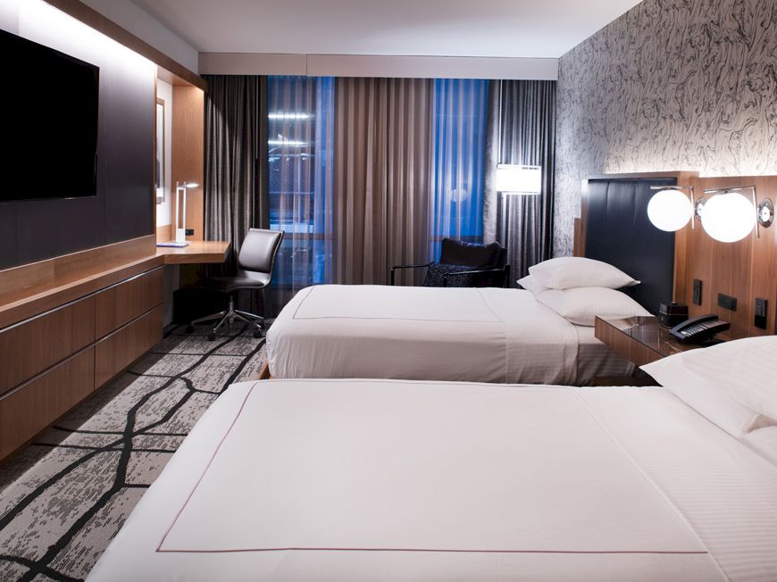 This image shows a modern hotel room with two neatly-made double beds, a desk with a chair, a TV on the wall, and contemporary decor.