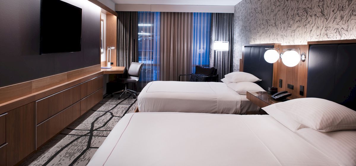 This image shows a modern hotel room with two neatly-made double beds, a desk with a chair, a TV on the wall, and contemporary decor.