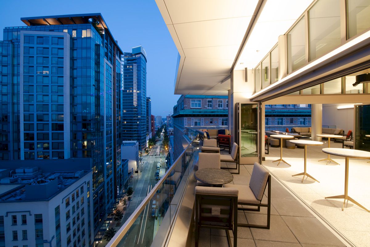 A modern balcony with seating and tables overlooks a cityscape with tall buildings at dusk, illuminated by city lights along the street.