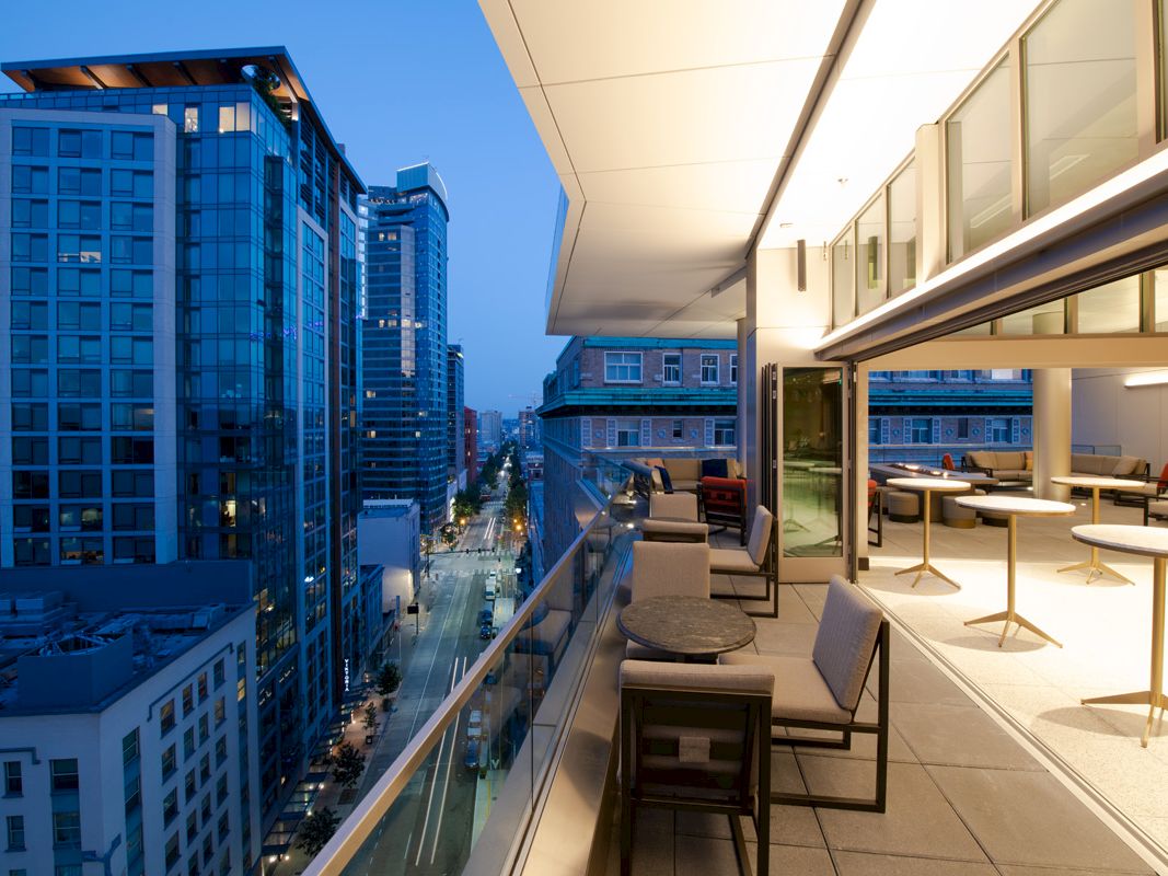 A modern balcony with seating and tables overlooks a cityscape with tall buildings at dusk, illuminated by city lights along the street.