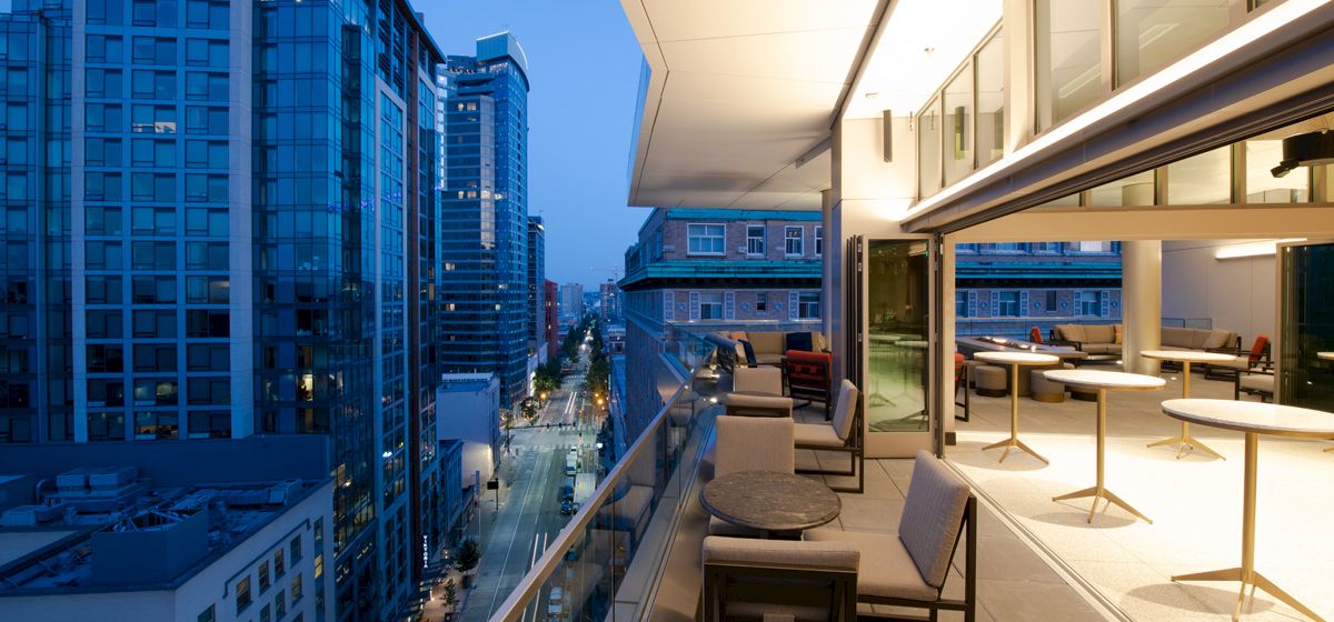A modern balcony with seating and tables overlooks a cityscape with tall buildings at dusk, illuminated by city lights along the street.