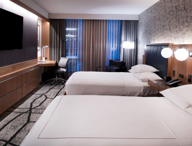 The image shows a modern hotel room with two neatly made beds, a flat-screen TV, a desk with a chair, and stylish decor.