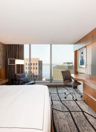 The image shows a modern hotel room with a bed, a desk, a chair, a large TV, and a view of city buildings through a large window.