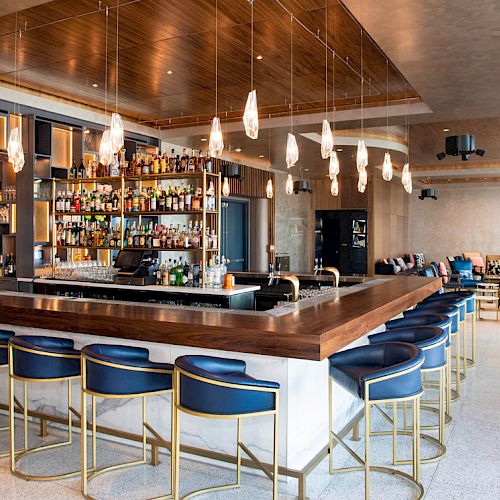 A stylish bar area with blue chairs, modern lighting, and well-stocked shelves, with a cozy seating area in the background.