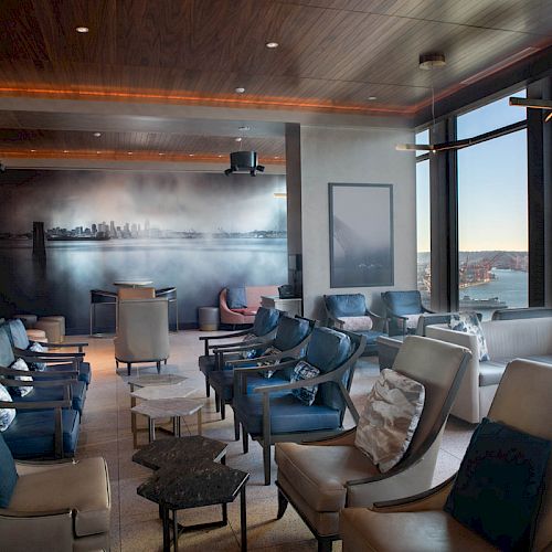 A modern lounge area with multiple chairs and tables, large windows offering a scenic waterfront view, ambient lighting, and wall art.