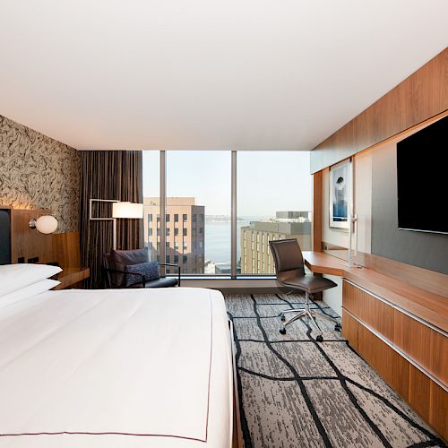 A modern hotel room featuring a large bed, TV, desk, chair, and a view of buildings through a floor-to-ceiling window ends the sentence.