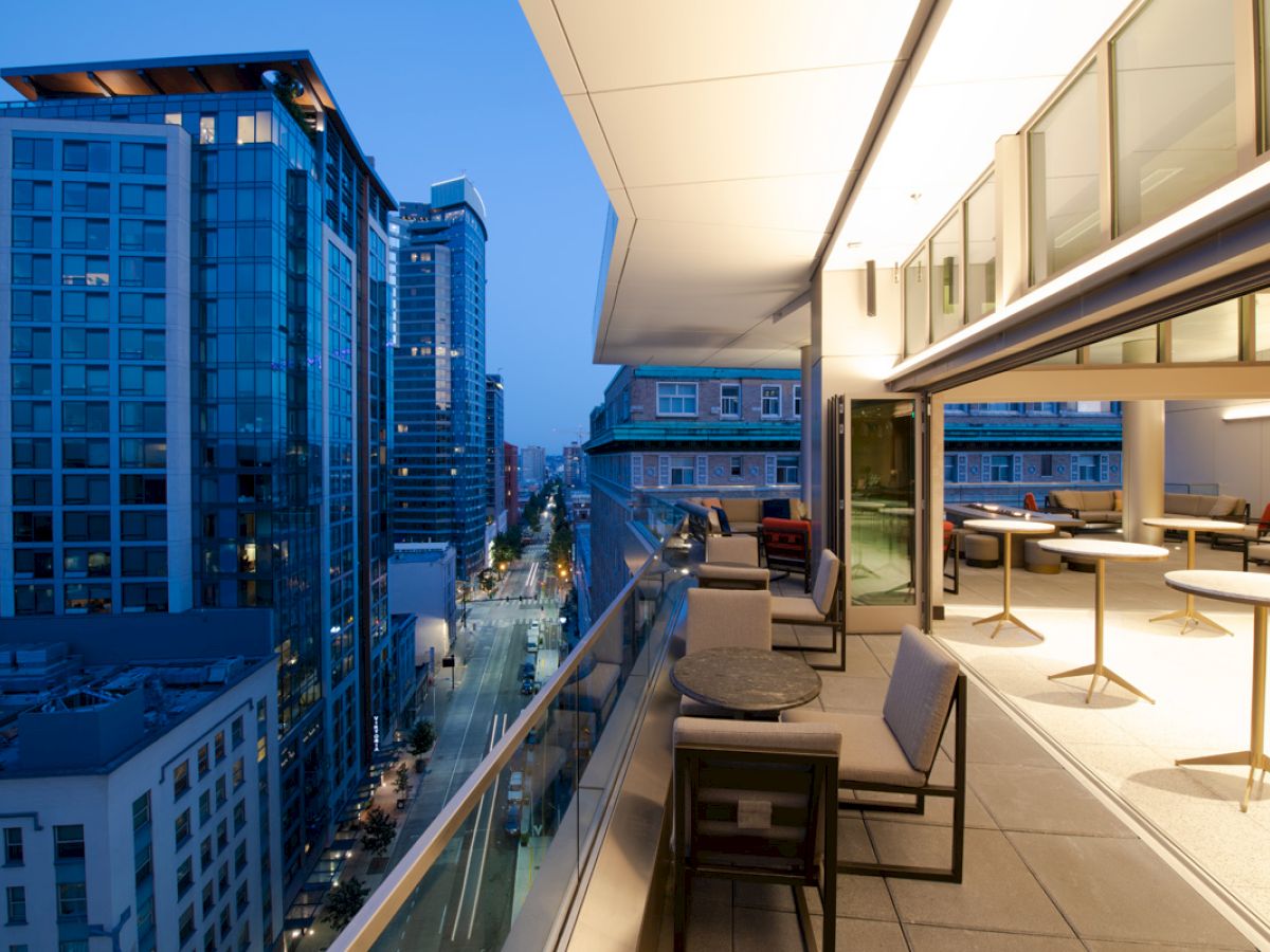 A modern cityscape during twilight with a view from a balcony featuring outdoor seating and buildings with glass facades lit up.