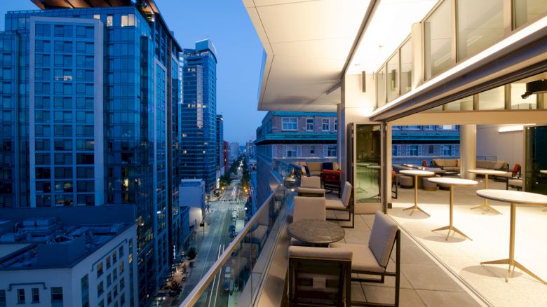 A modern cityscape during twilight with a view from a balcony featuring outdoor seating and buildings with glass facades lit up.