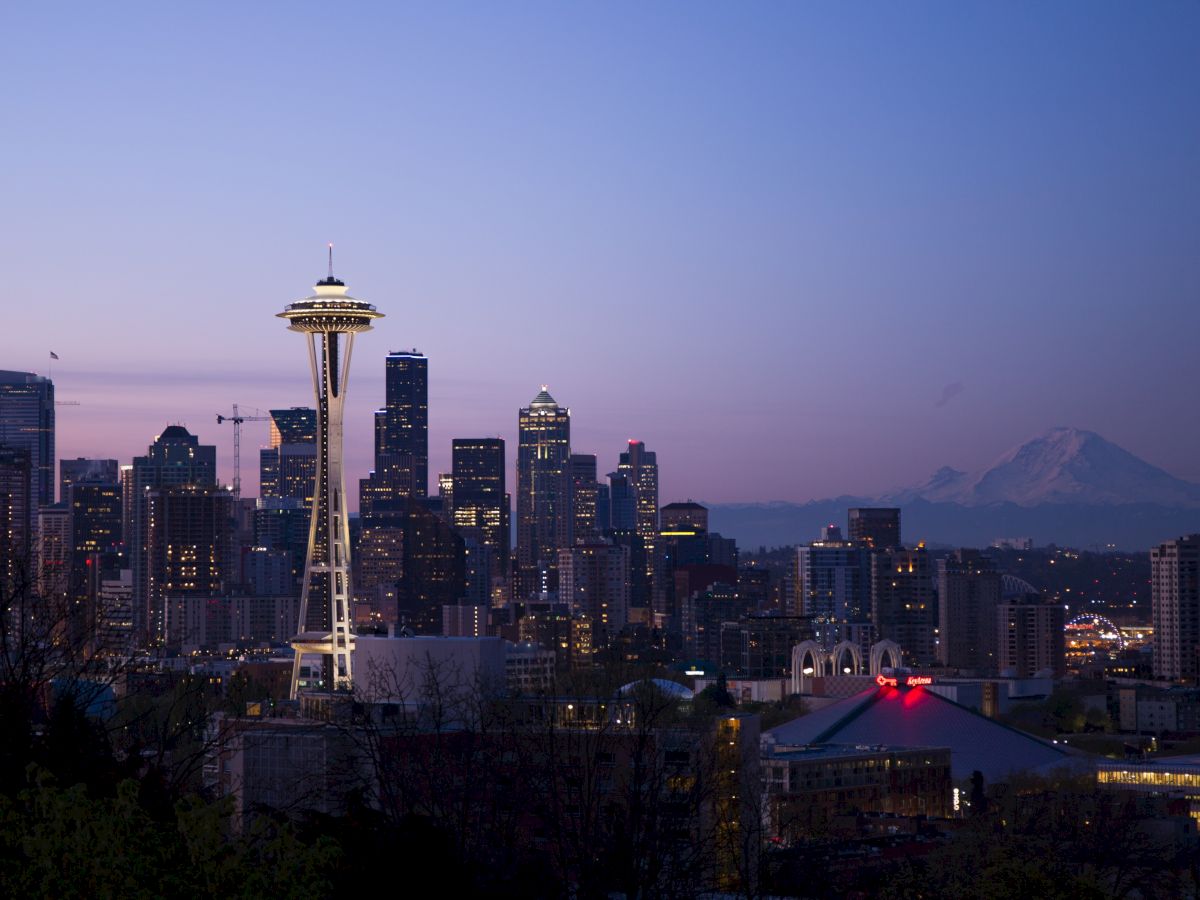 The image shows the Seattle skyline at dusk, featuring the Space Needle prominently in the foreground with Mount Rainier in the background.
