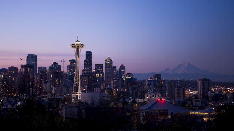 The image shows the Seattle skyline at dusk, featuring the Space Needle prominently in the foreground with Mount Rainier in the background.