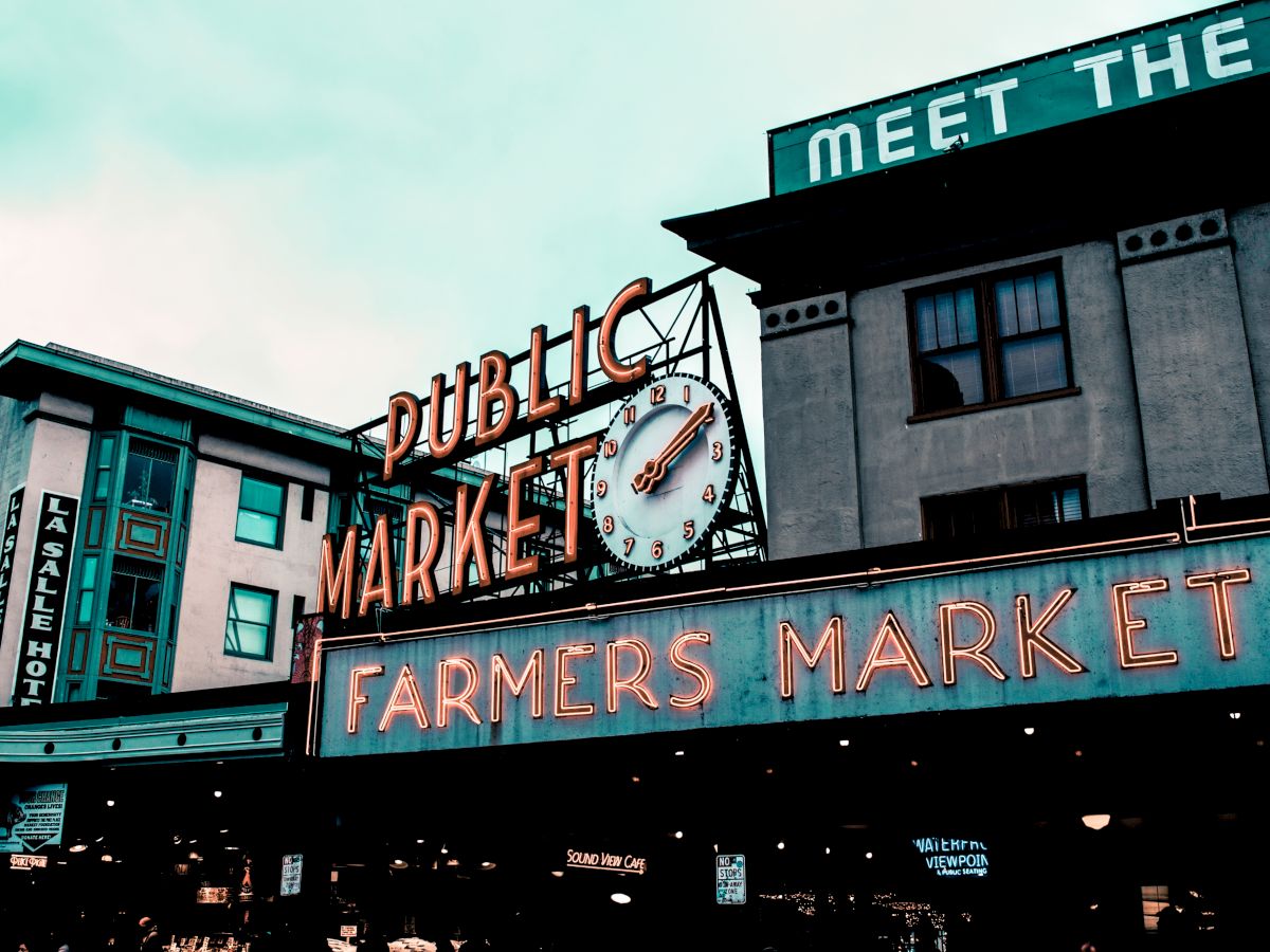 The image shows a public market with a prominent sign reading "Farmers Market" in neon lights. There is also a clock and surrounding buildings.