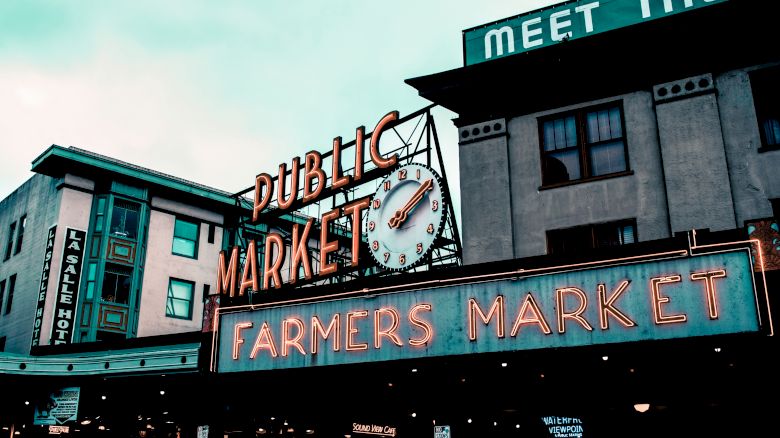 The image shows a public market with a prominent sign reading "Farmers Market" in neon lights. There is also a clock and surrounding buildings.