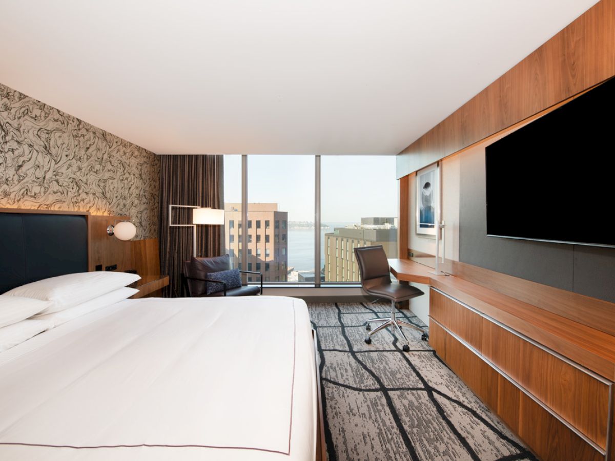A modern hotel room featuring a king-sized bed, large TV, desk with a chair, and a view of city buildings through the window.