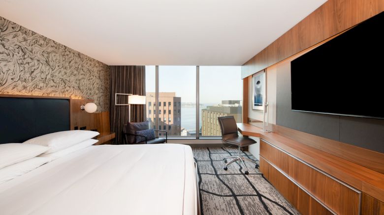A modern hotel room featuring a king-sized bed, large TV, desk with a chair, and a view of city buildings through the window.