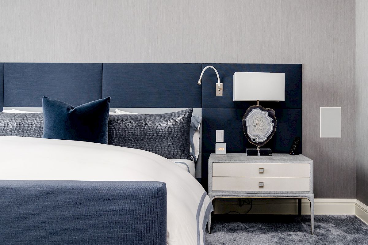 A modern bedroom featuring a bed with blue and gray bedding, a white nightstand with a lamp, and a plush carpet.