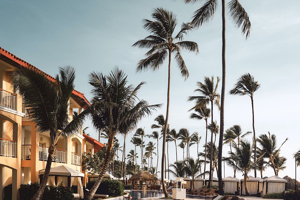 The image shows a serene tropical resort with palm trees, a pool, lounge chairs, and a multi-story building in the background under a clear sky.