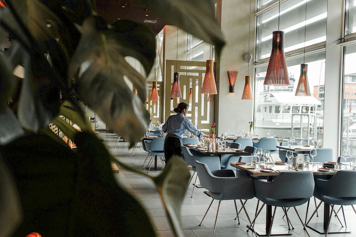 A person is setting tables in a modern restaurant with plants in the foreground and stylish lighting fixtures overhead.