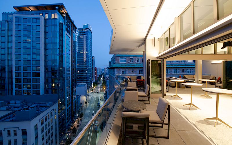A modern cityscape at dusk featuring high-rise buildings and a stylish rooftop terrace with tables and chairs overlooking the street below.