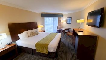 This image shows a hotel room featuring a large bed with white sheets and a green runner, two nightstands, a desk and chair, a dresser with a TV.