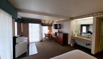 A hotel room with a bed, a desk, a TV, a dresser, a microwave, a mini-fridge, a sink, a jacuzzi, and a balcony with a view through glass doors.