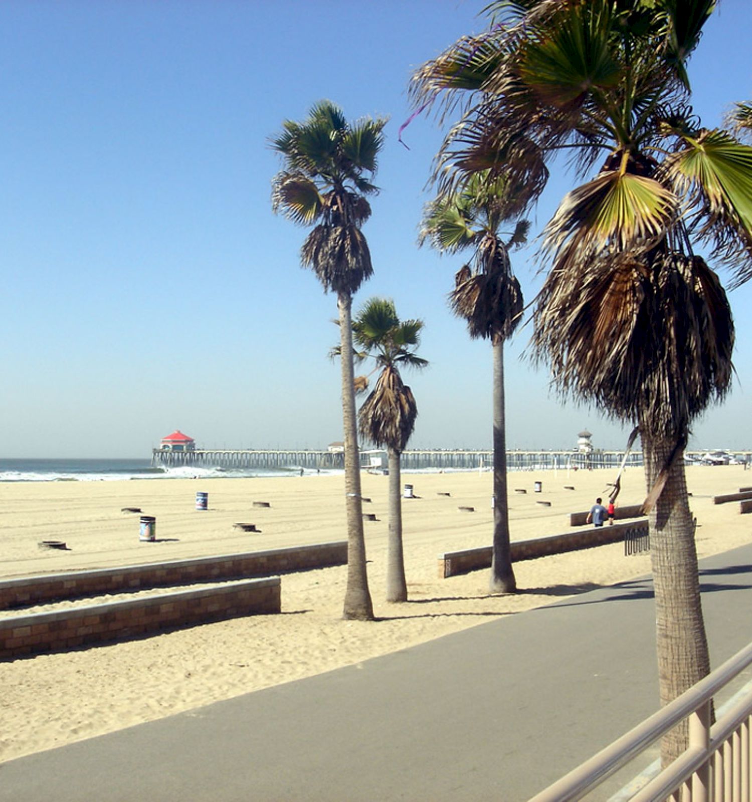 An empty beach with palm trees, a boardwalk, and a distant pier under a clear blue sky–a peaceful coastal scene.