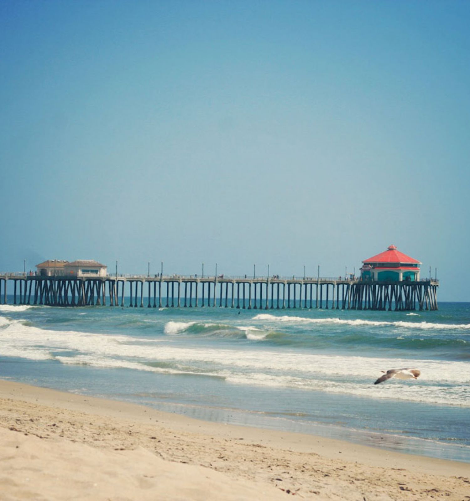 A beach scene with two people walking on the sand, waves crashing, and a long pier extending into the ocean, ending with a red-roofed structure.