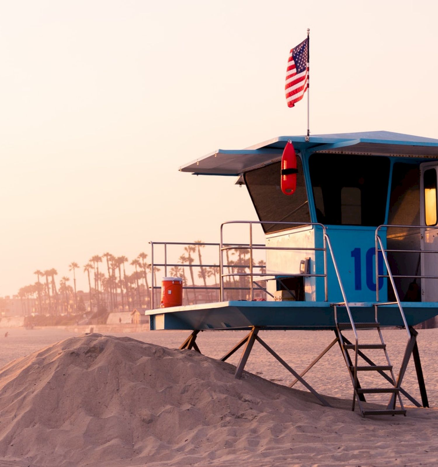 A lifeguard tower with the number 16, an American flag, safety equipment, and a sand mound on a beach during sunset, with palm trees in the background.