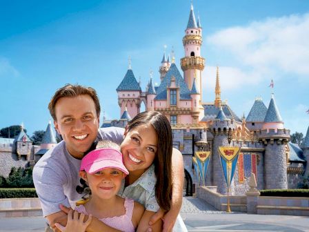 A family of three, two adults and a child, pose in front of a fairy tale castle. The child wears a pink hat, and everyone is smiling.