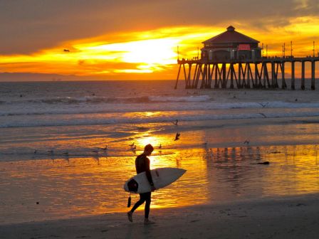 A surfer walks on the beach at sunset, with a pier extending into the ocean and the sky illuminated in orange and yellow tones.