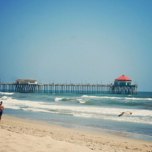 Two people walk on a sandy beach with a long pier and a red-roofed structure in the background, beside a gently rolling ocean.